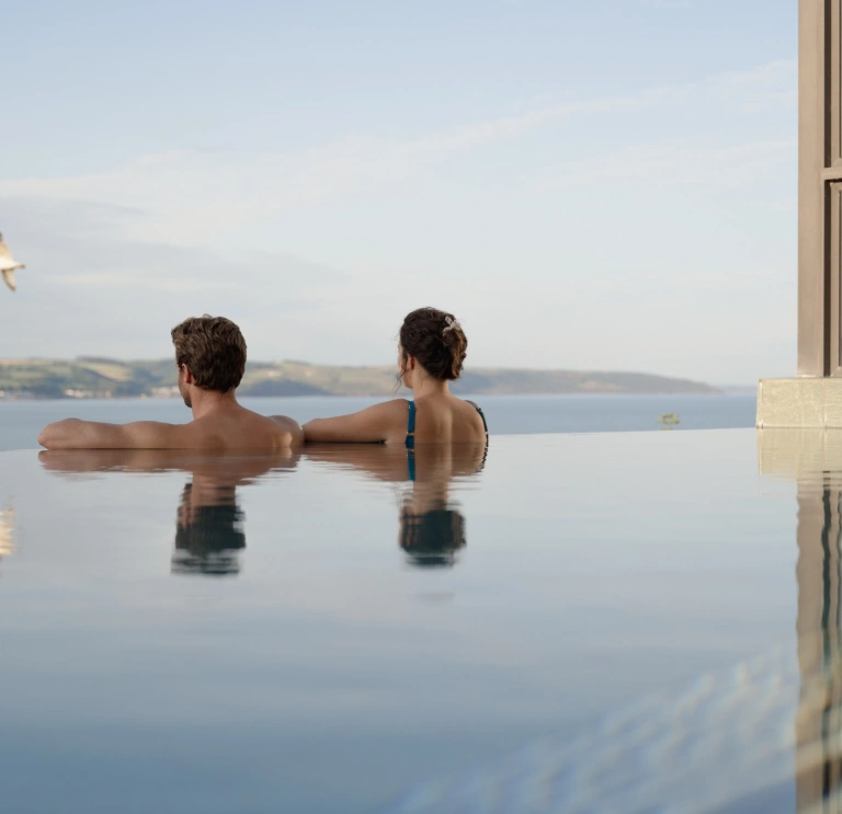 two people in an infinity pool looking out at the view of a headland of green trees.