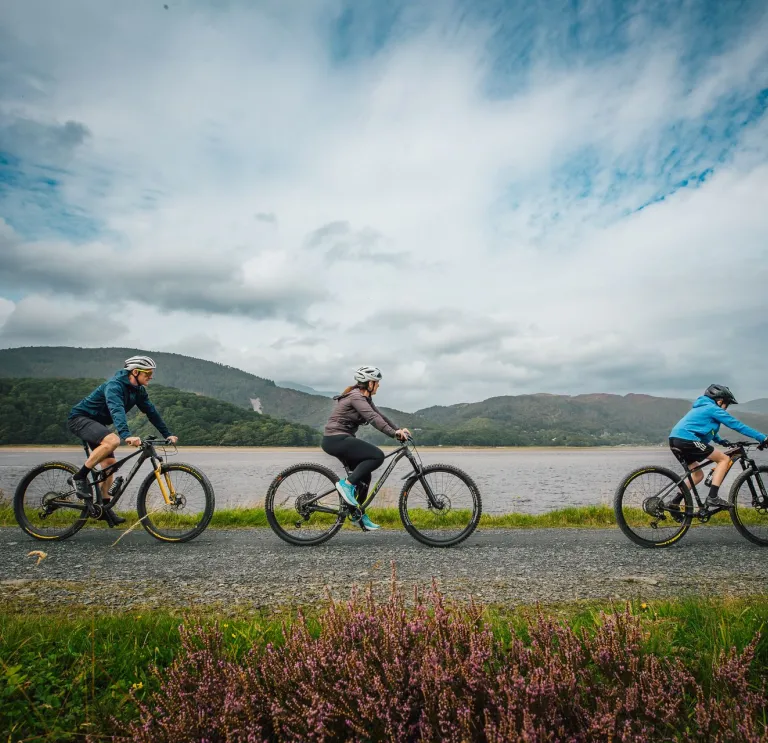 Three cyclists on a riverside pathway, with mountains in the background.