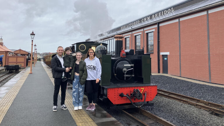 A family of two adults and two children standing by a small green steam engine at a station.