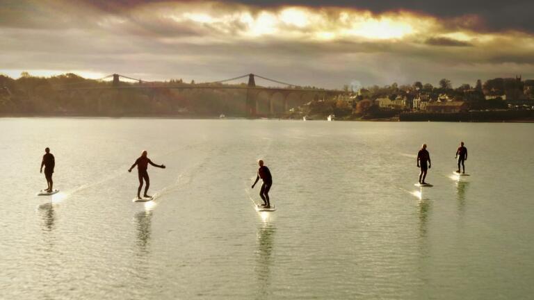 Five people riding efoil boards over a stretch of water at sunset.