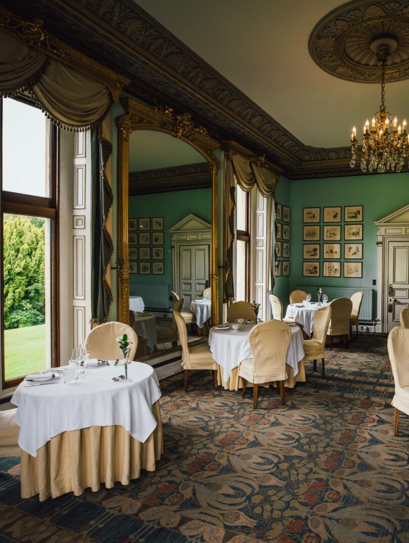 An ornate dining room in a Victorian mansion. Pale green walls, round tables with plates and silverware, windows looking out over a green lawn. 