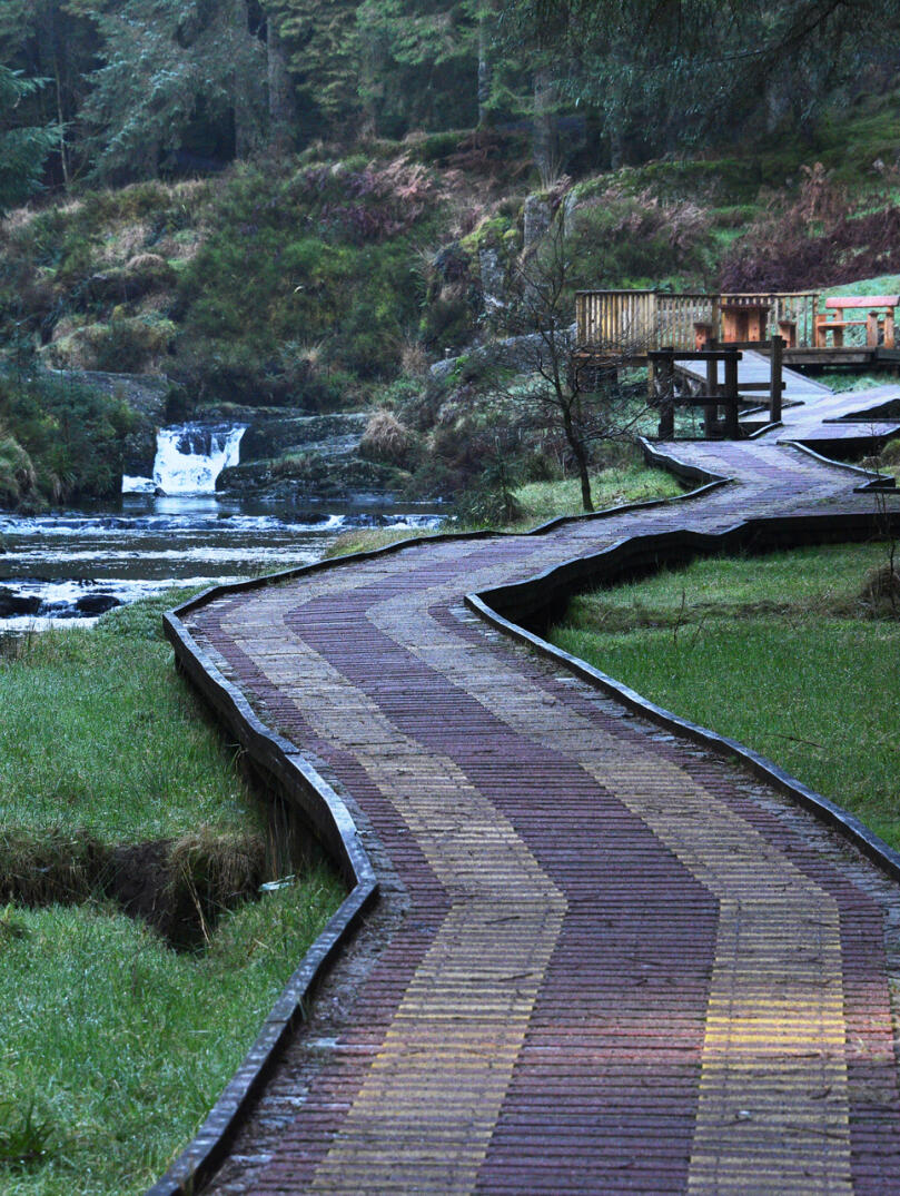 A wide boardwalk in woodland leading to a viewing point, alongside a river.