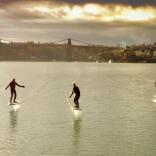 Five people riding efoil boards over a stretch of water at sunset.