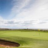 A golf course with well-maintained green fairway under a clear blue sky. A sand bunker is visible in the foreground, and a yellow flag marks the hole on the green. Two golfers can be seen in the distance against a backdrop of coastal terrain.