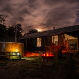 Nachtaufnahme einer Glamping-Hütte mit Whirlpool und beleuchtetem Feuerplatz im Freien. 