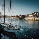 A wide river with a sailing boat in the foreground and a quay in the background.