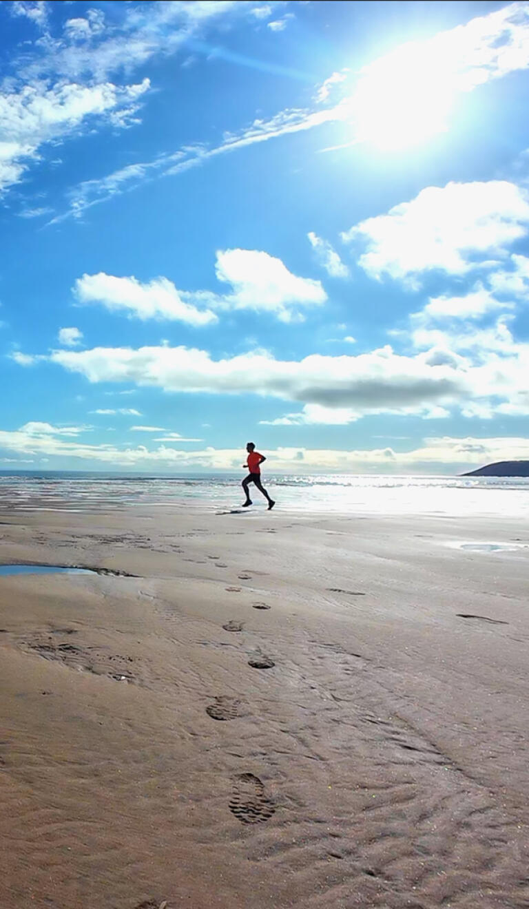 A man in tshirt and shorts running along a wide sandy beach, under blue skies.