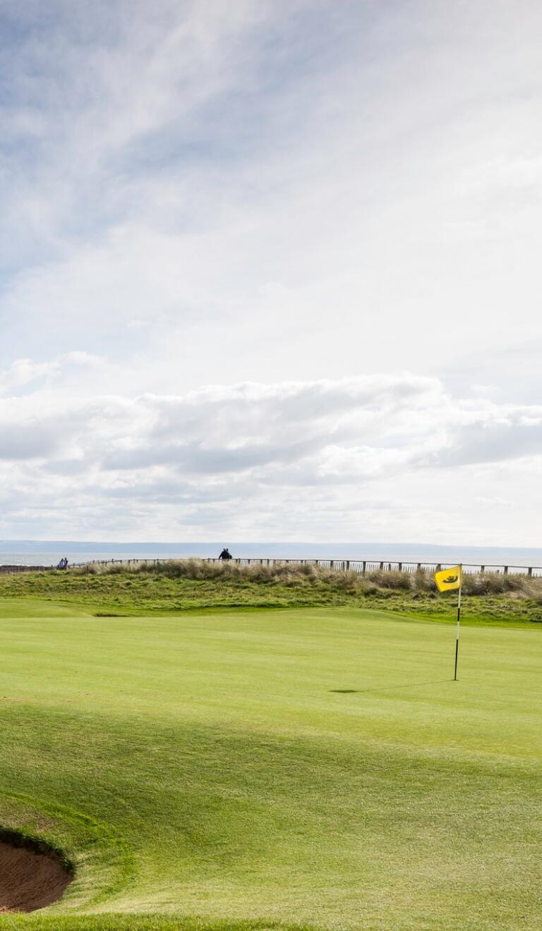 A golf course with well-maintained green fairway under a clear blue sky. A sand bunker is visible in the foreground, and a yellow flag marks the hole on the green. Two golfers can be seen in the distance against a backdrop of coastal terrain.