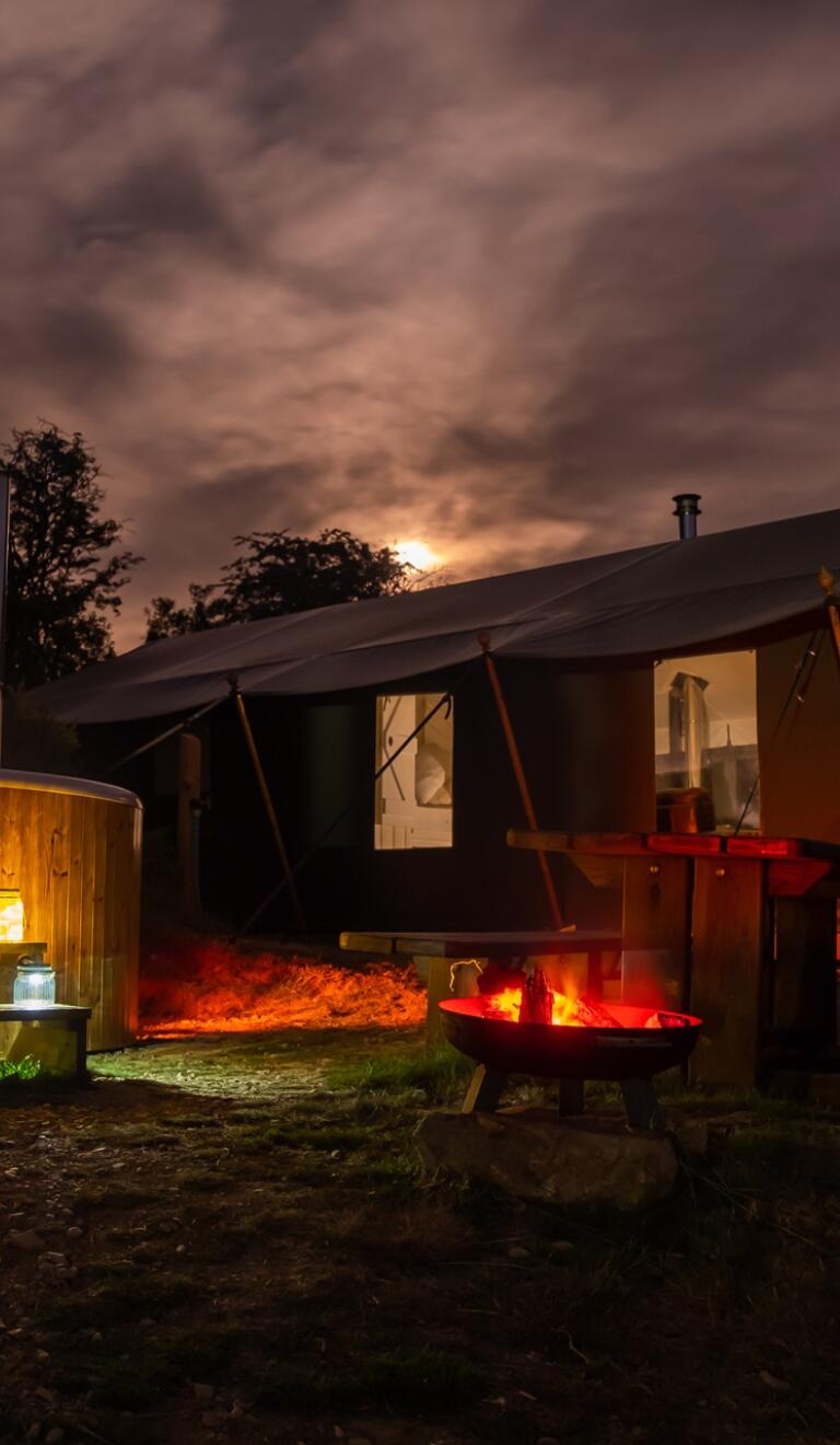 Nachtaufnahme einer Glamping-Hütte mit Whirlpool und beleuchtetem Feuerplatz im Freien. 